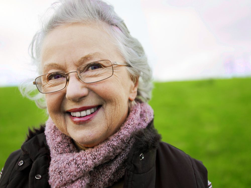 An elderly woman in glasses smiles in a field